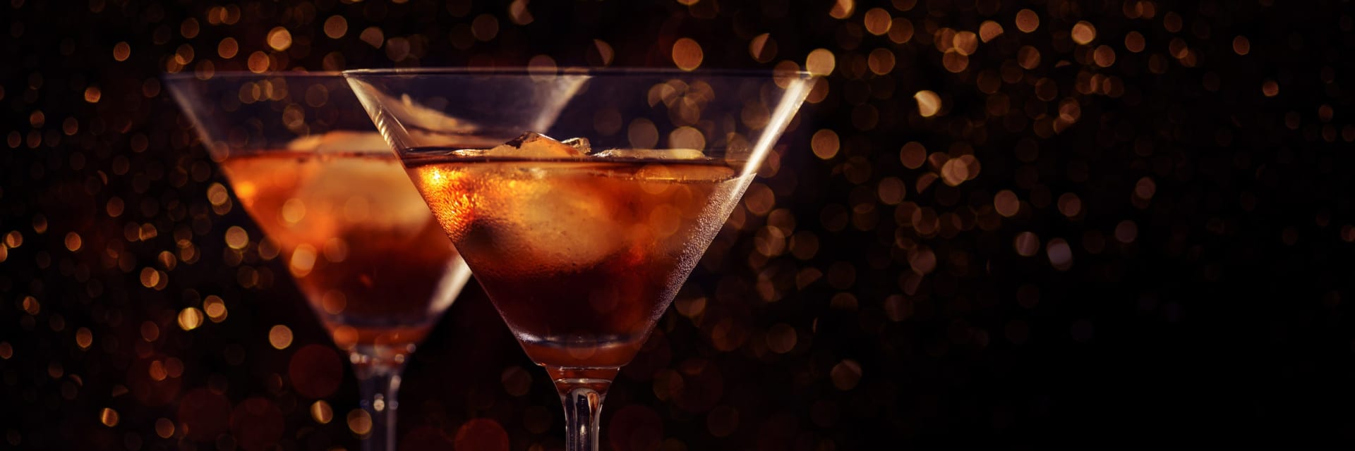 Two martini glasses filled with amber-colored drinks and ice, set against a dark background with warm golden bokeh lights, creating a festive and elegant atmosphere.