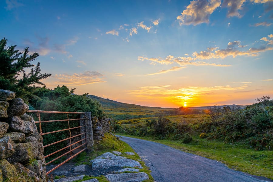 A scenic countryside road winds through green fields and shrubs at sunset, with a stone wall and metal gate on the left, and a colorful sky filled with clouds and sunlight.