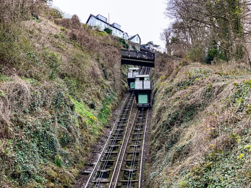 A funicular railway with two green and white carriages travels up and down steep parallel tracks, surrounded by greenery and hills, passing under a bridge with houses above.